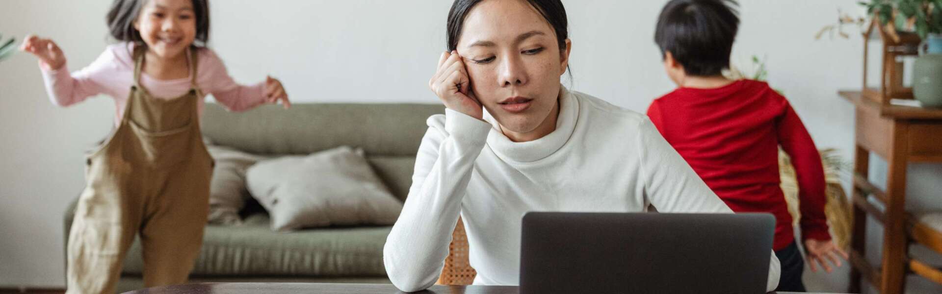 A tired-looking mother sits at a laptop while two children run in circles behind her.