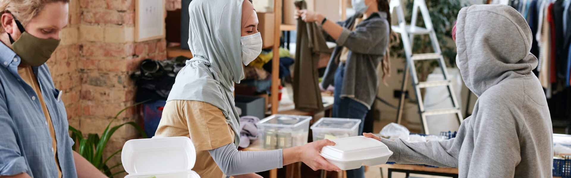 Two people wearing masks fill takeout containers and pass one to a person wearing a hoodie