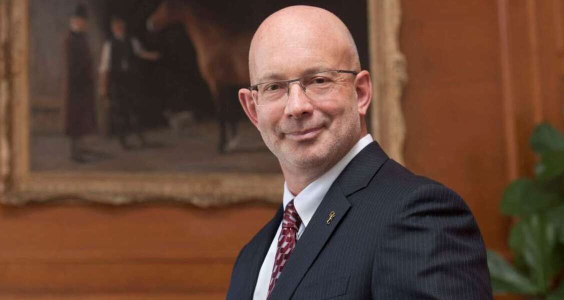 A person in a black suit, white shirt and red tie with wire frame glasses smiles in front of a painting of a horse on a wood panelled wall.