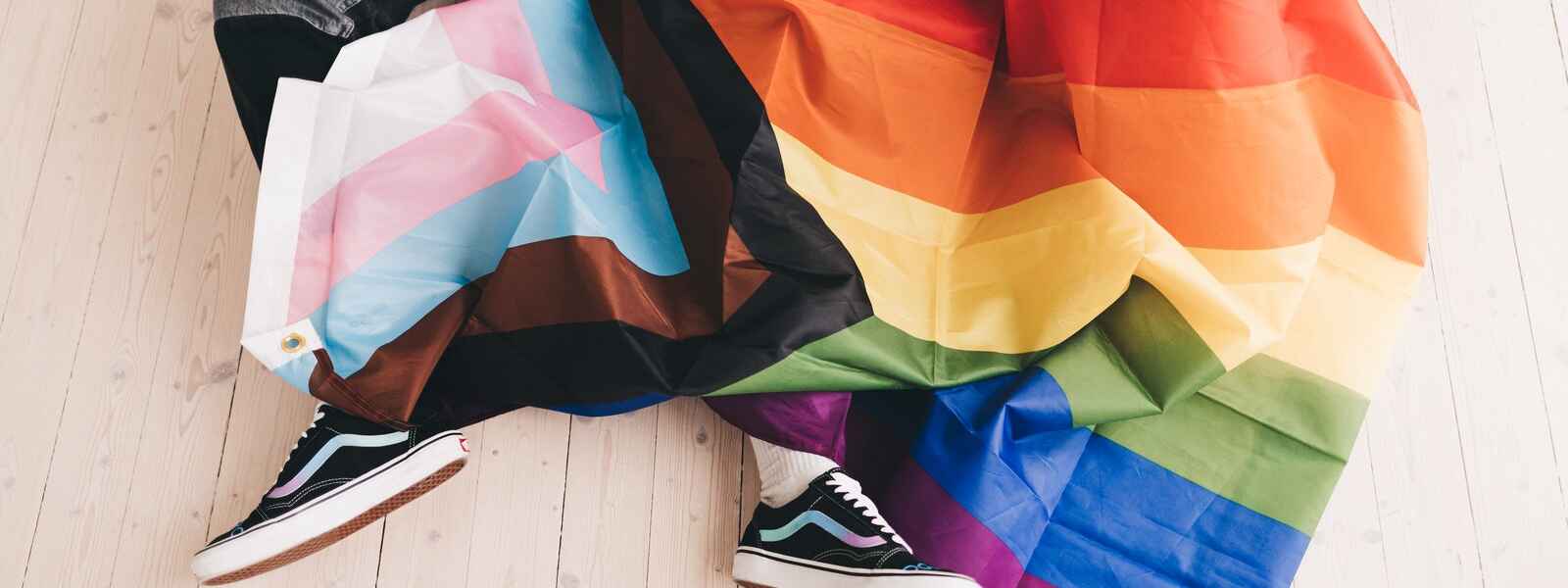 Two people sit with Progress Pride flag draped over them.