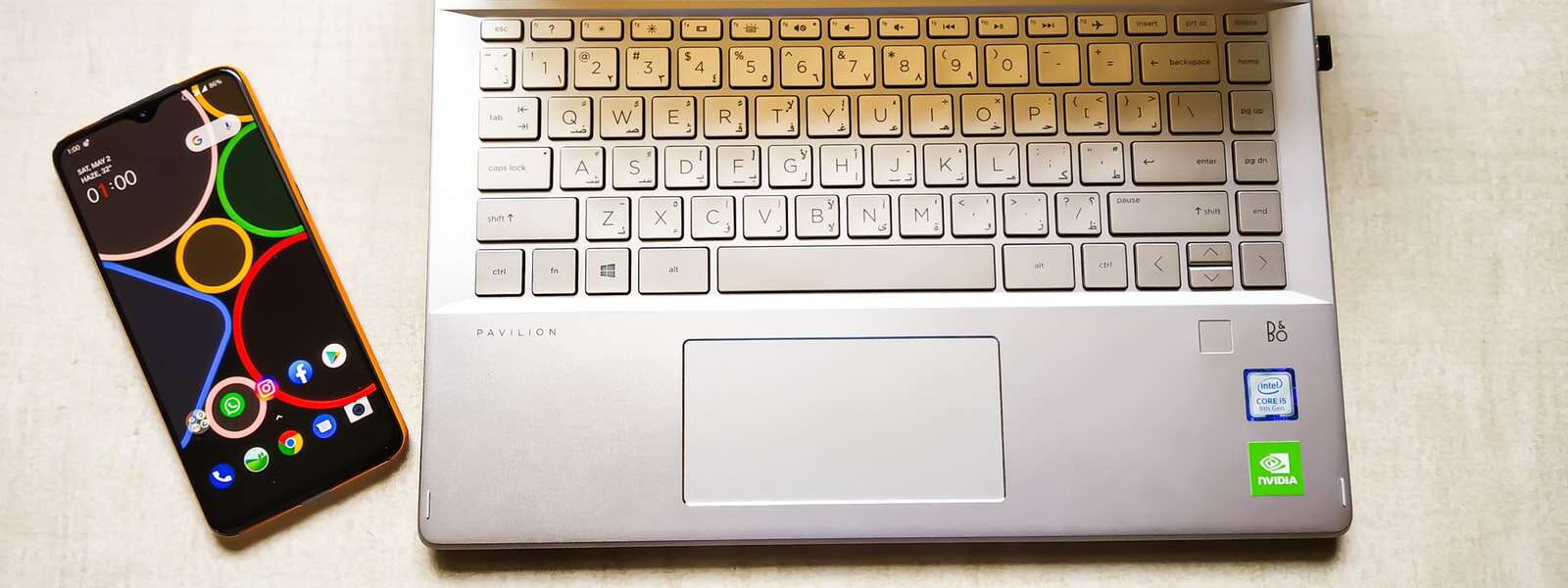 A phone set to its homescreen sits next to an open laptop on a table.