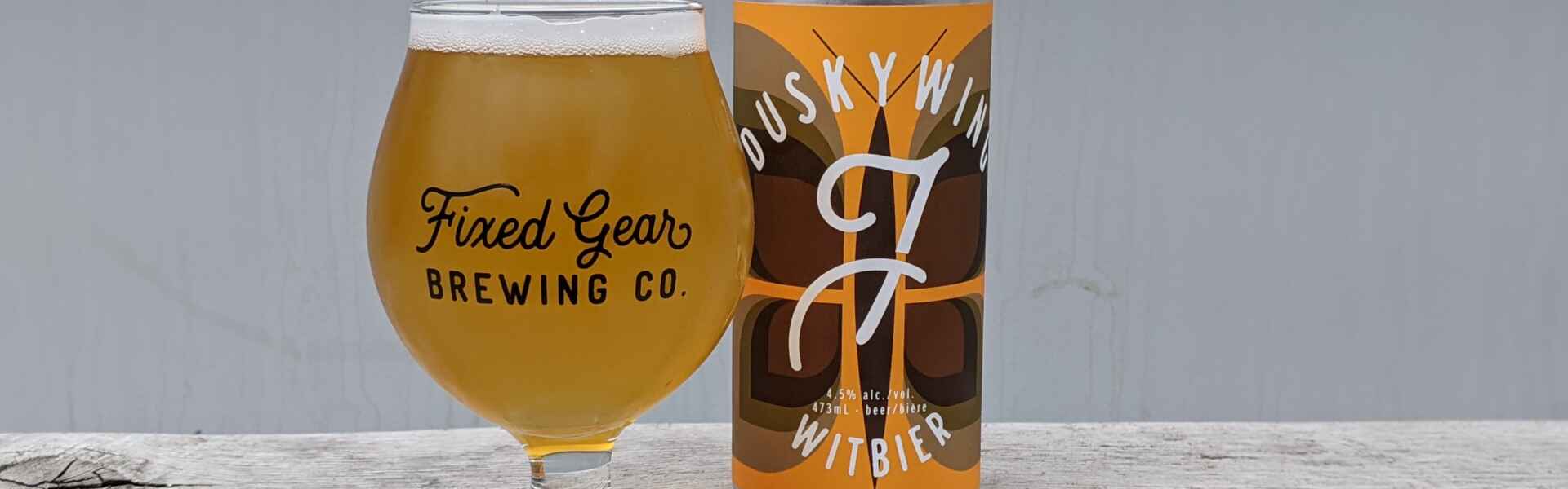 A glass of a yellow beer sits on a wooden table next to a silver can with a brown butterfly on an orange label.