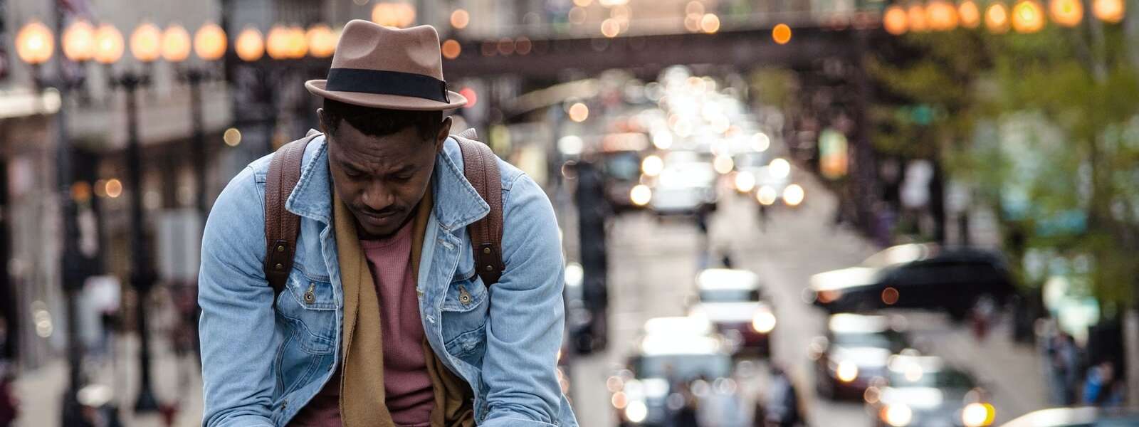A man in a leather jacket looking down while sitting on a ledge in a city