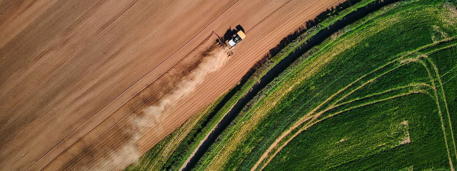 An aerial view of fields, which spilt the image in half diagonally across the centre. In the top half a tractor drives through a brown field. In the bottom half is a green field.