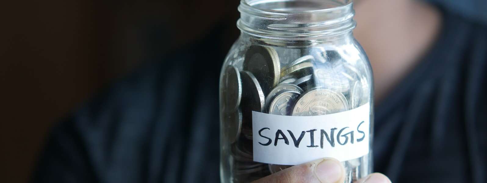 a hand holds a glass jar filled with coins labelled 'Savings'