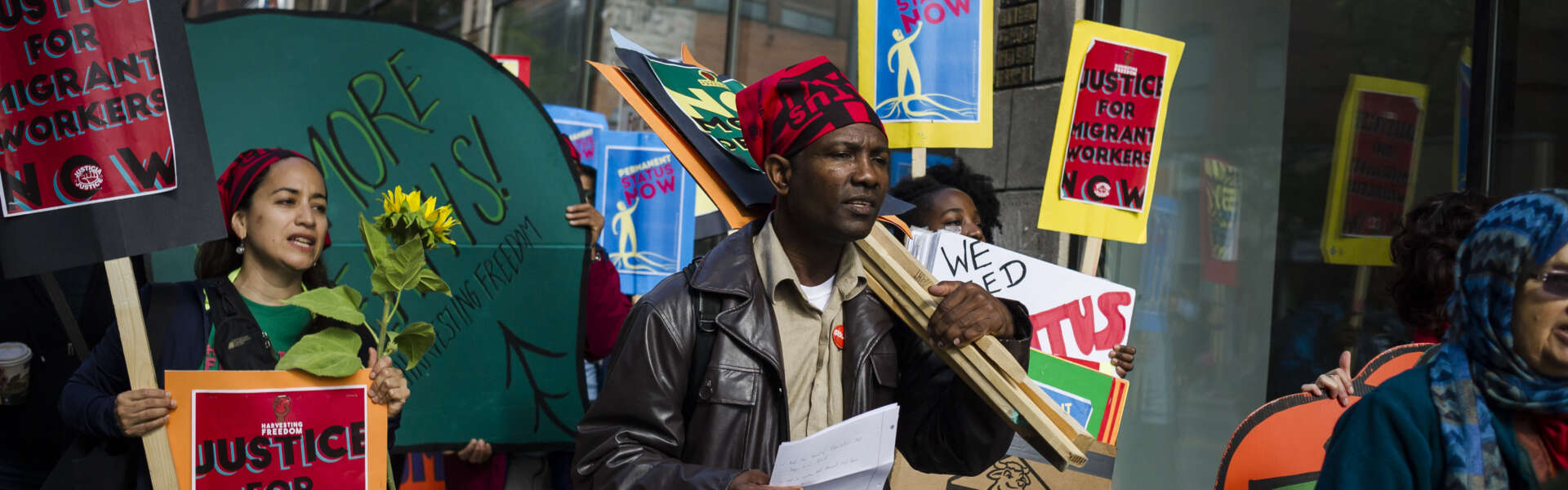 A man in brown clothes, leather jacket and red head scarf carries protest sign with crowd of people.