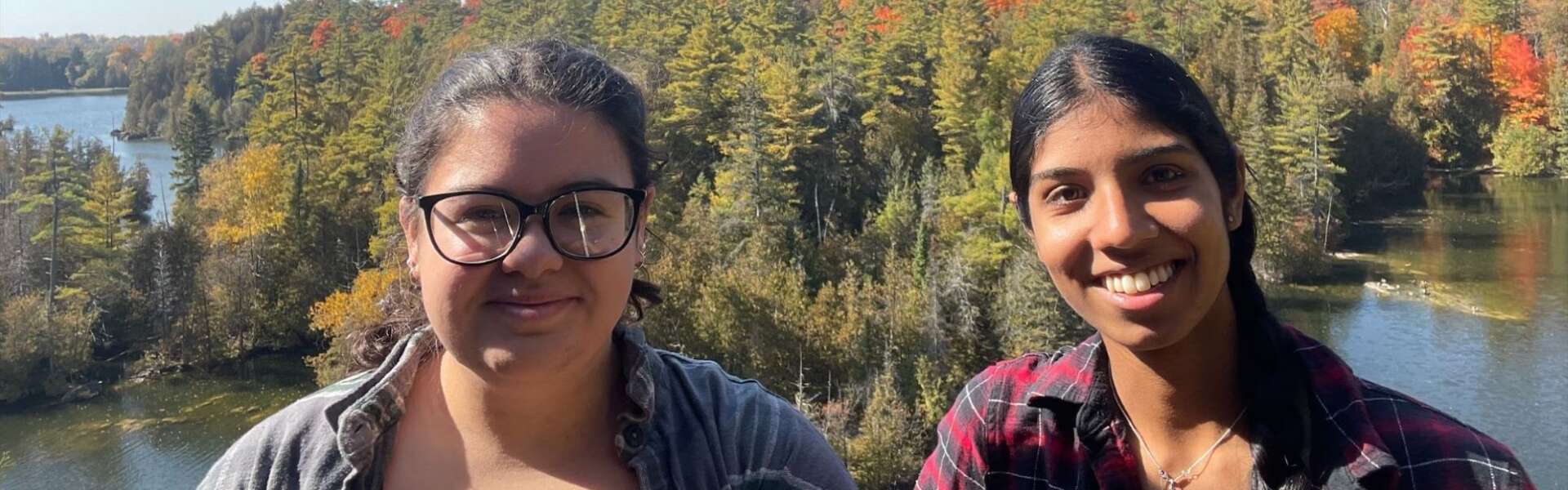 Julie-Ann Arbour (left) and Gurleen Chana take a selfie in front of lakes and a forest in the fall.