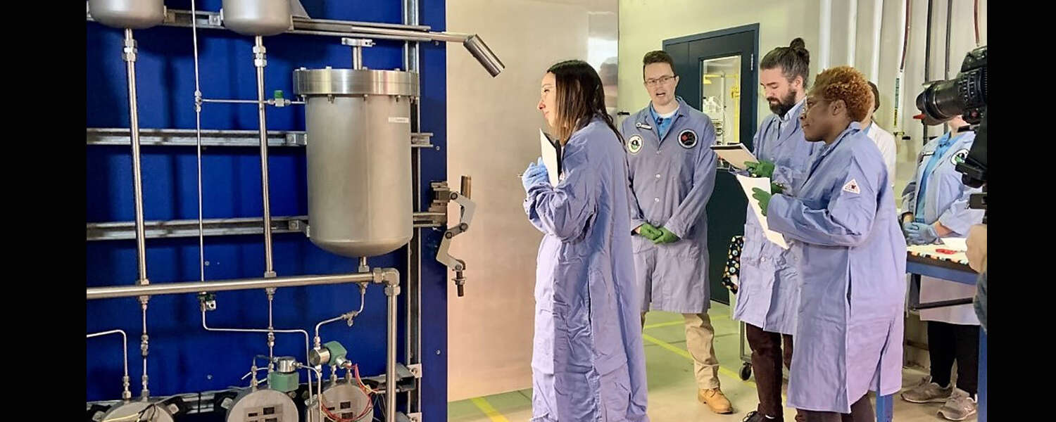 Four people in lab coats look inside a large blue chamber featuring water filtration pipes on the side