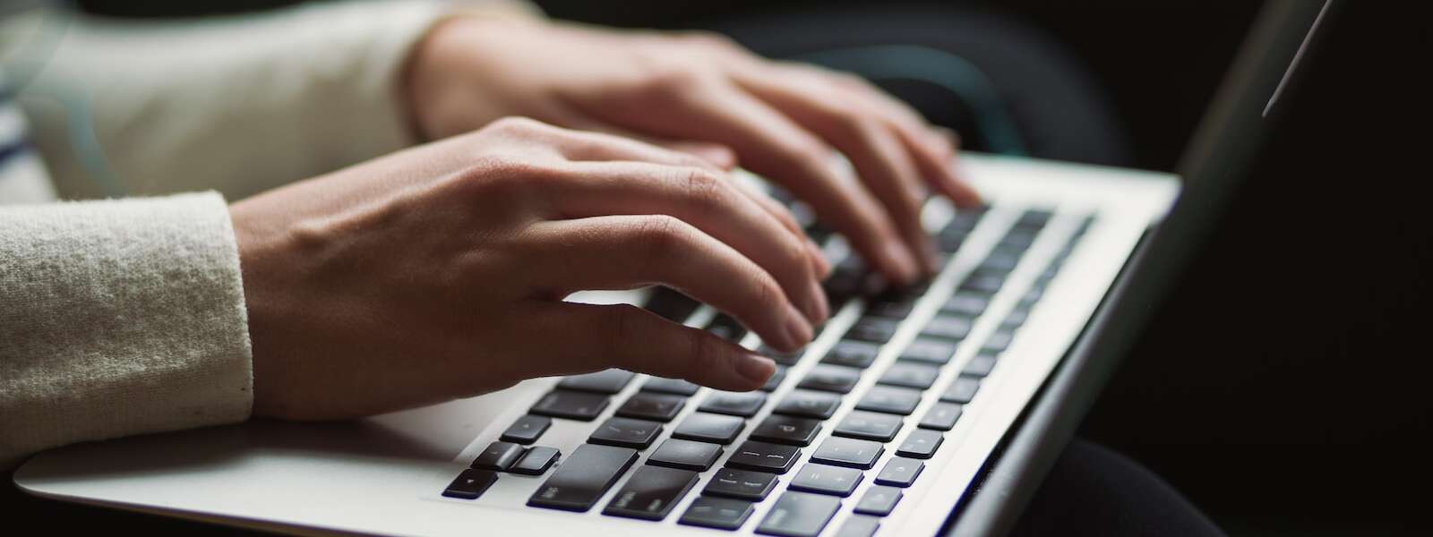A person wearing a white sweater types on a black keyboard on a pale gray laptop