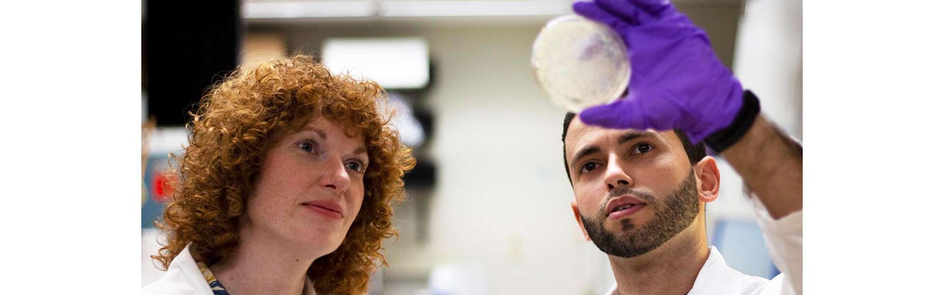 Two people in lab coats look at a petri dish held up to the light by a gloved hand