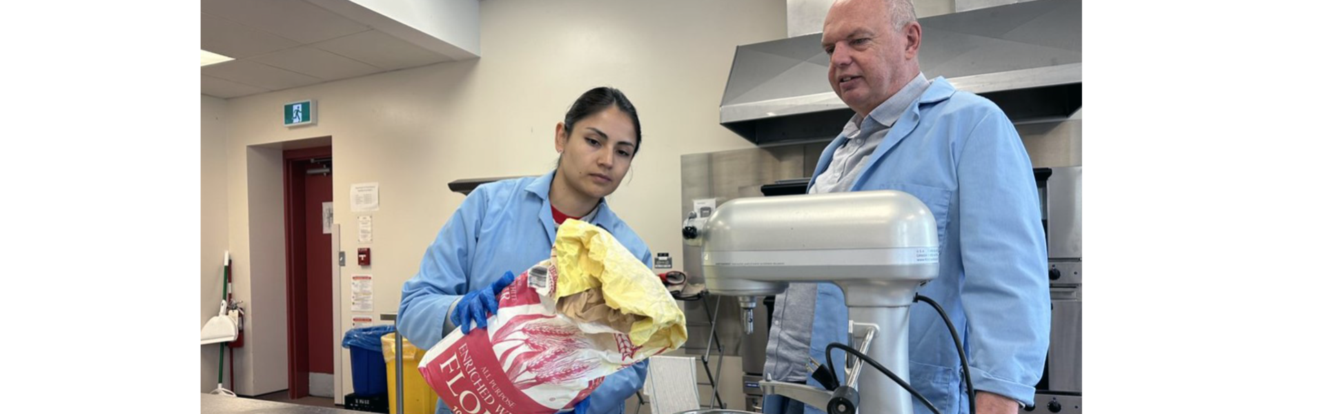 Two people wearing lab coats prepare to pour a bag of flour into a large stand mixer