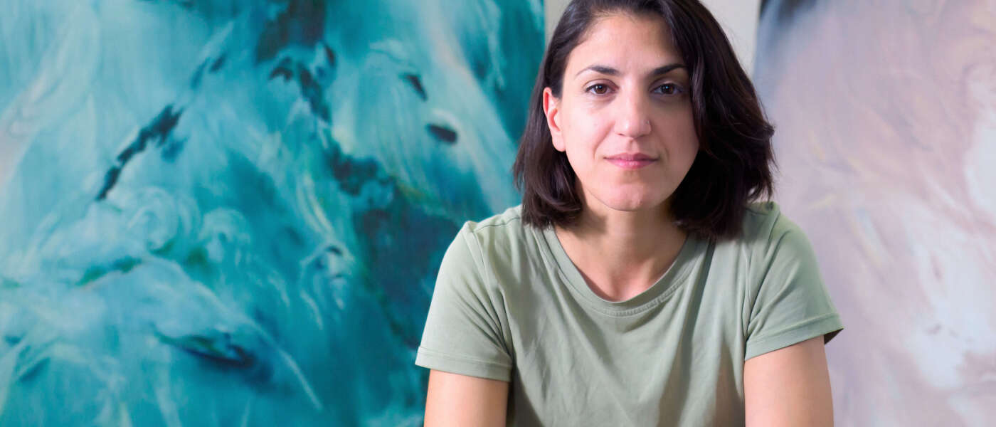 A person with shoulder-length dark brown hair sits with their elbows resting on their bent knees wearing a light green top and dark green pants in front of their blue and green painting.
