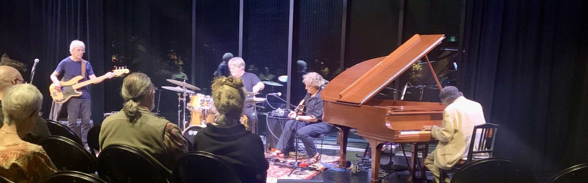 Four musicians including one on a grand piano play in a dark theatre with large windows behind