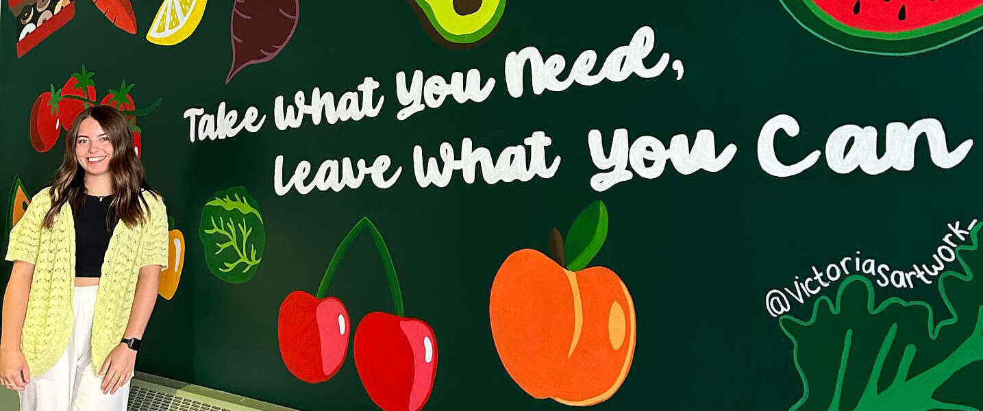 A person with long, wavy brown hair stands in a black top, yellow sweater, khaki pants and white shoes in front of a mural of red, orange, green and yellow fruits and vegetables with white text reading, take what you need, leave what you can.