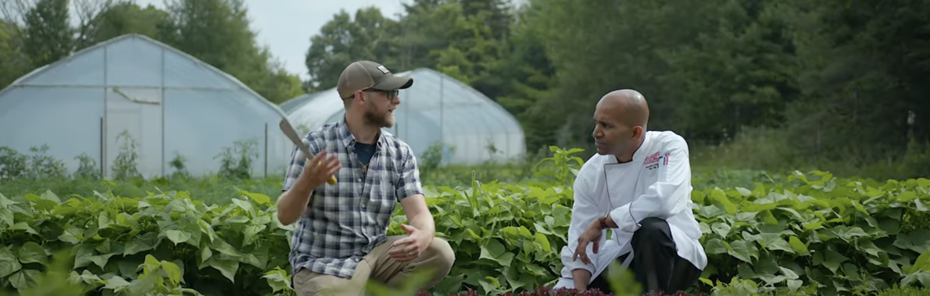 A man in a plaid shirt, a beard, glasses and a baseball cap holds a small shovel while crouched in a garden beside a second man dressed in chef whites.