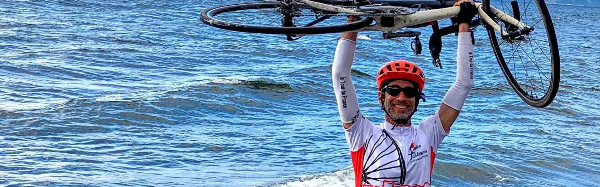 A man in red, white and black cycling clothes raises his bike over his helmeted head standing on the shore of the ocean.