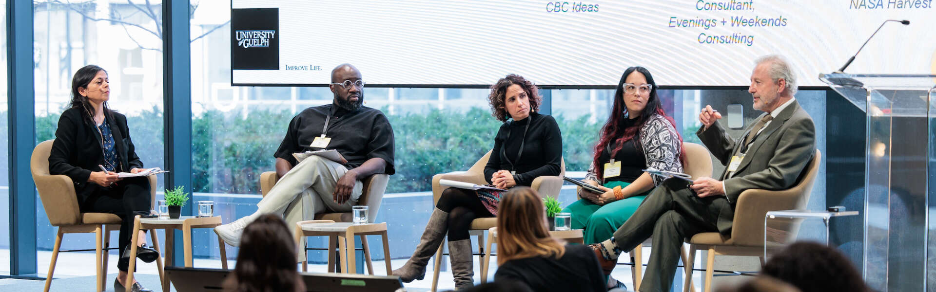 Five people set in chairs in front of a large screen while speaking to a room of participants
