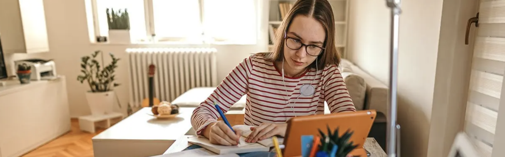 a woman with earbuds sits at a desk in an apartment taking notes with a pen while watching a tablet