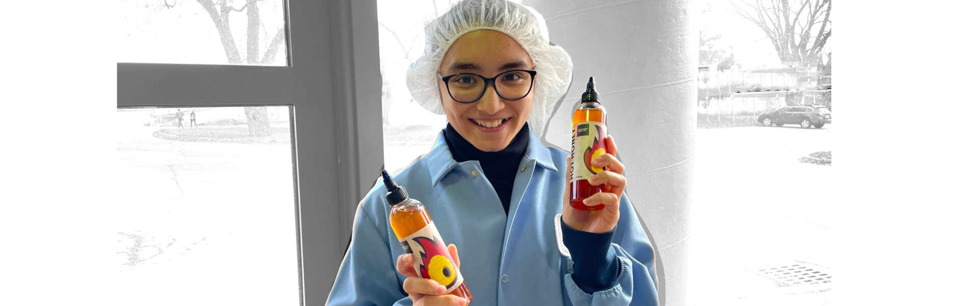 a person in a lab coat and hair net smiles while holding two bottles of honey