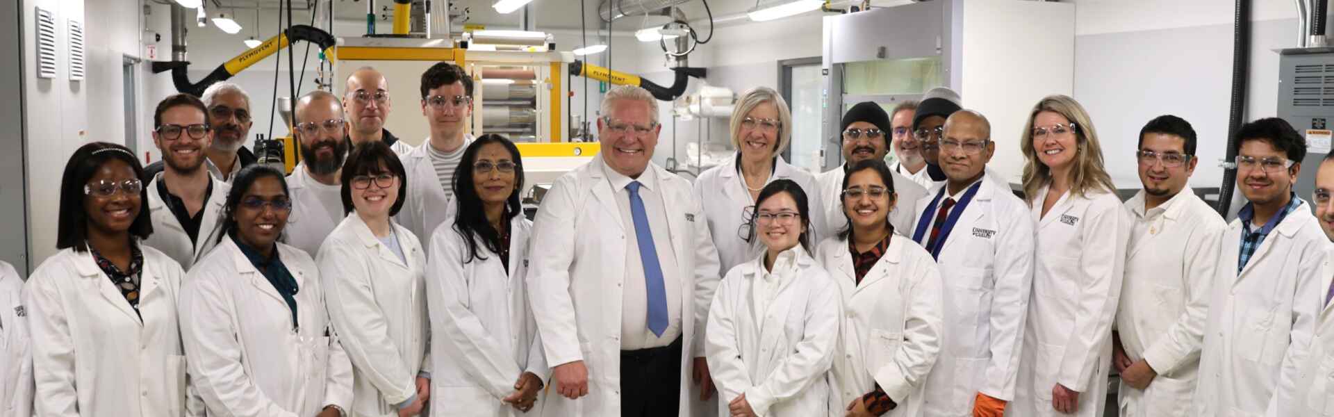 A group of 20 people in lab coats pose for a portrait while standing in a machinery area