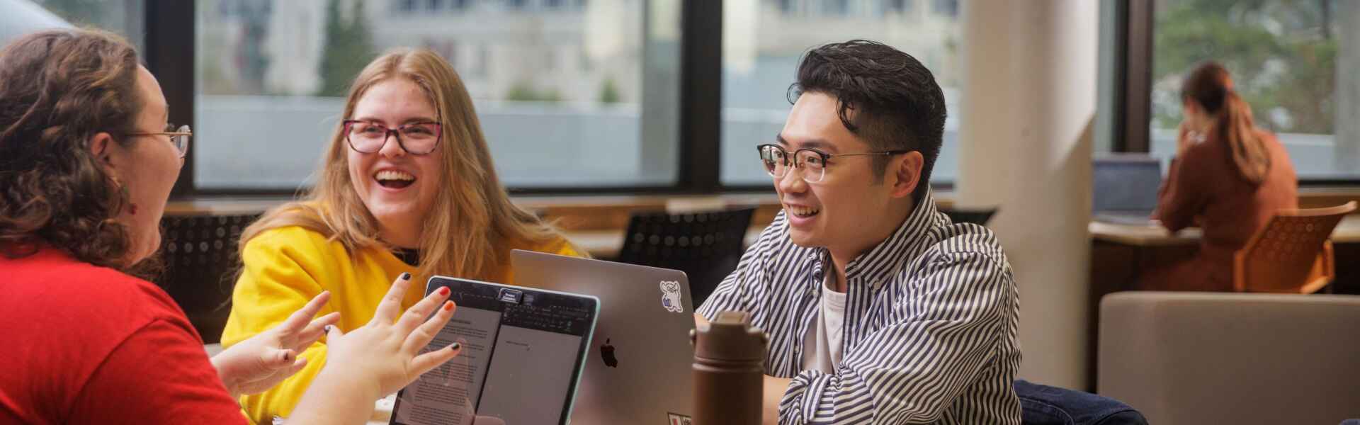 Two people sitting at a desk with laptops before them laugh while a third person at the desk gesticulates while speaking