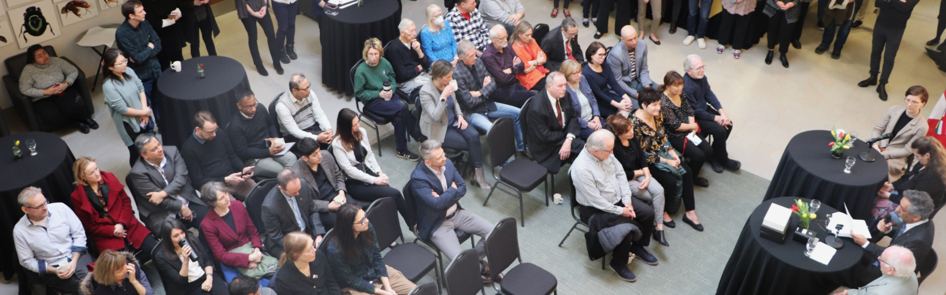 View from second floor of the recognition event in the Centre for Biodiversity Genomics. Roughly 50 attendees sit in chairs in the middle while another 50 stand around the perimeter. Dr. Paul Hebert and panel sit at round high tables at the front of the room.