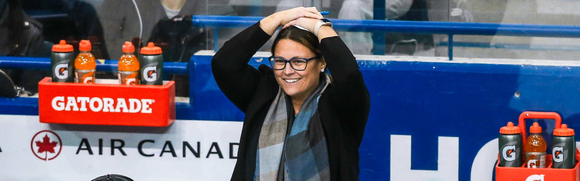 A person in a black coat and blue scarf stands with their hands on their head behind a bench full of hockey players in blue uniforms.
