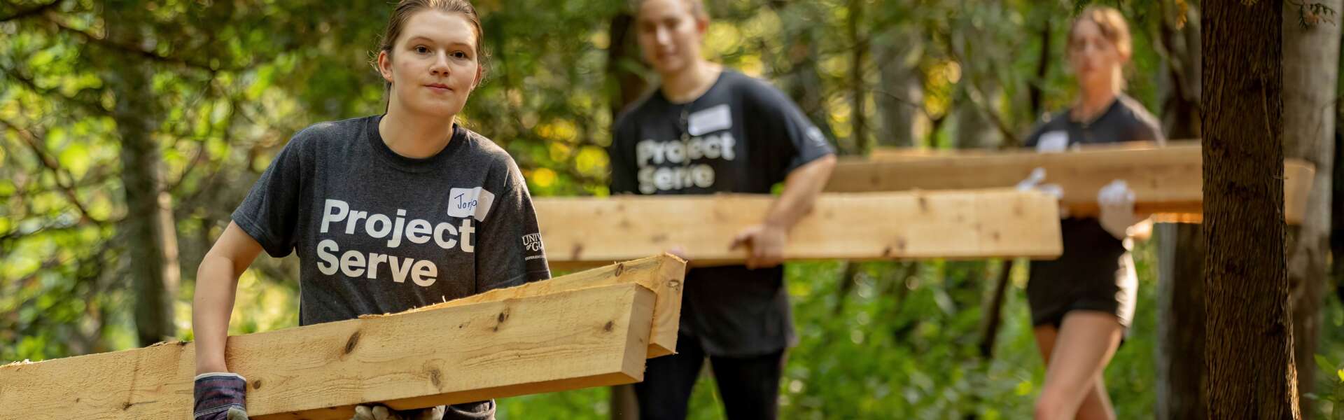 Students wearing project serve tshirts carry long wooden planks through a wooded trail.