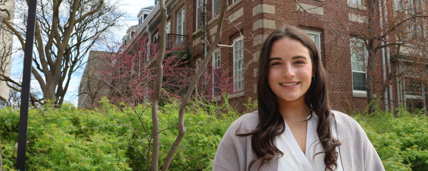 A student with long brown hair, wearing a white blouse and a grey cardigan stands smiling in front of a red brick building and landscaped greenery on campus.
