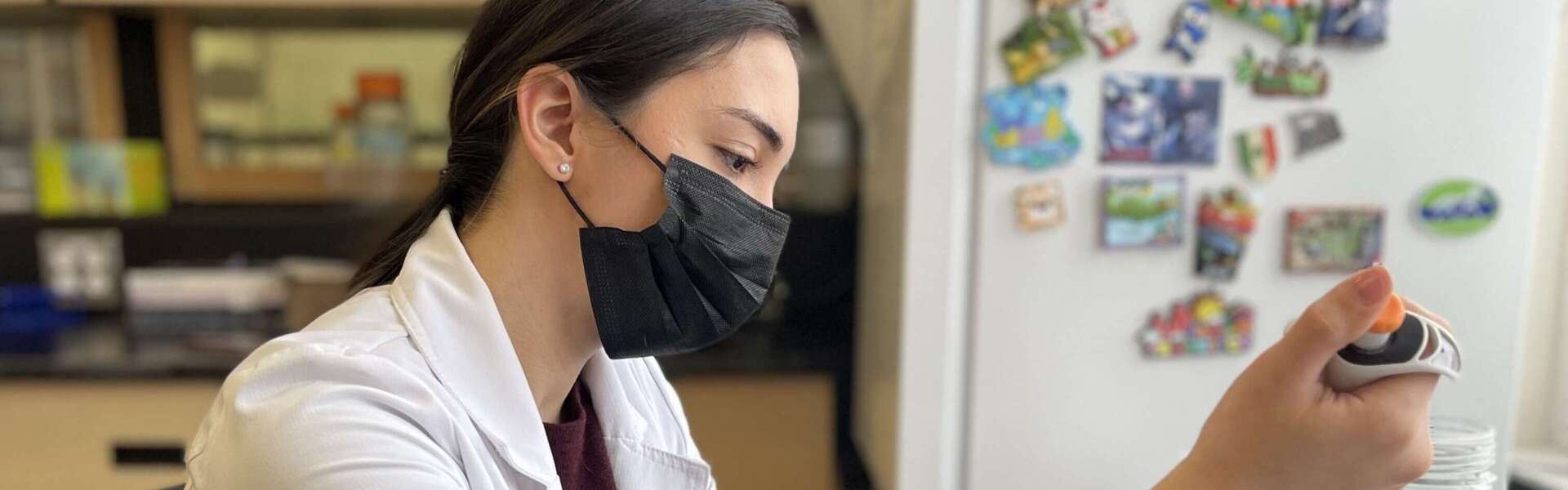A person with brown hair wearing a black face mask and a white lab coat sits at a desk and transfers specimen.