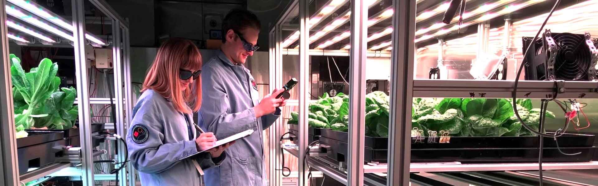 two students in lab coats and sunglasses examine plants on stands in a controlled environment growing facility