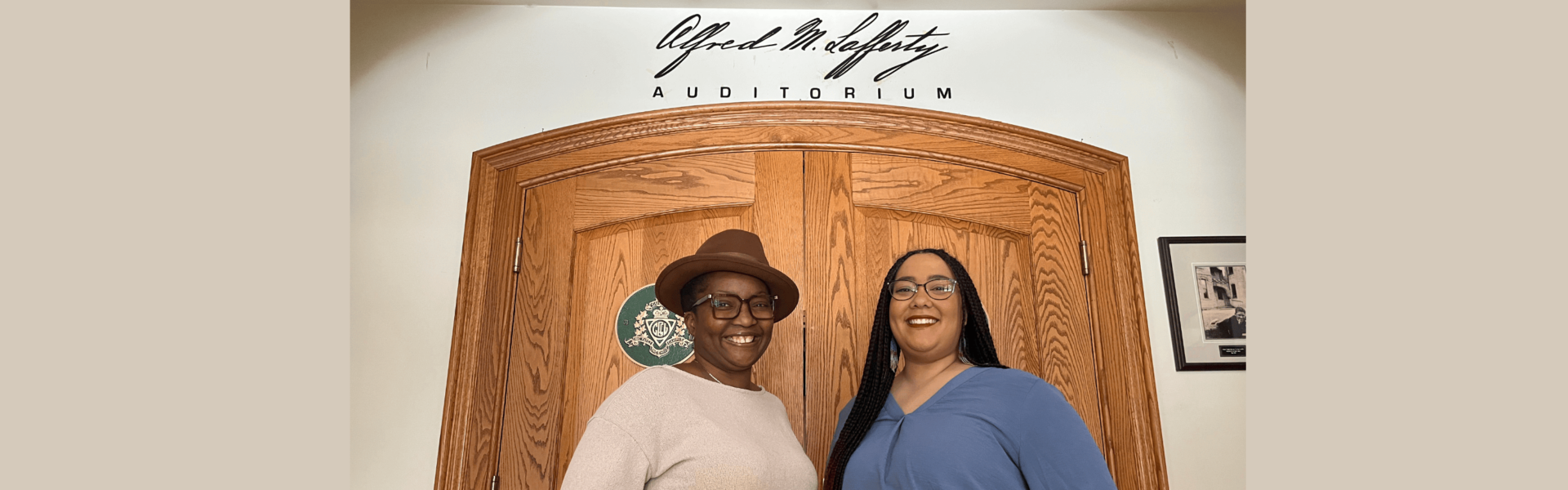 Two Black women stand facing one another and smiling, one in a beige sweater and brown hat and one in a blue shirt and black braids in front of two wooden doors with script on the wall above them.