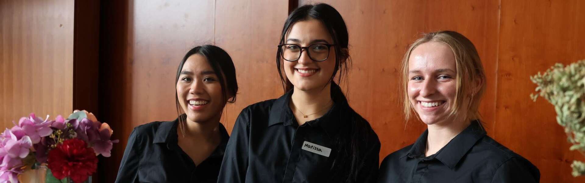 Three students in black button-up long sleeve shirts stand together smiling behind a counter with a vase of multicoloured flowers in front of a wooden wall.