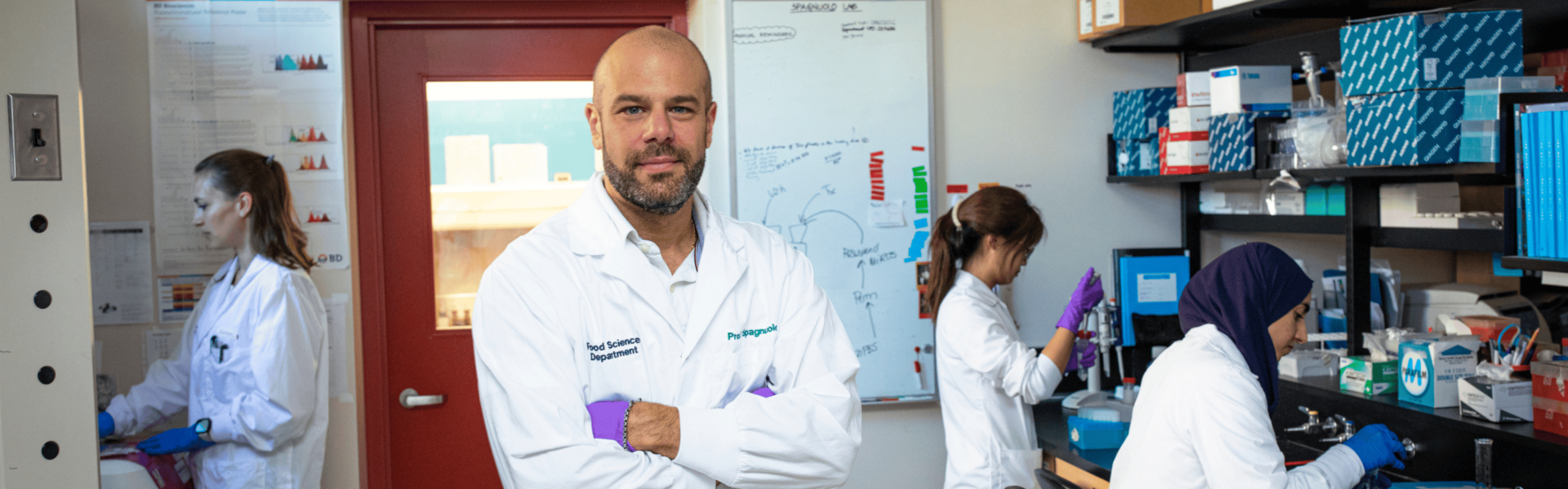 Dr. Paul Spagnuolo smiles at the camera as a few researchers work behind them in a lab