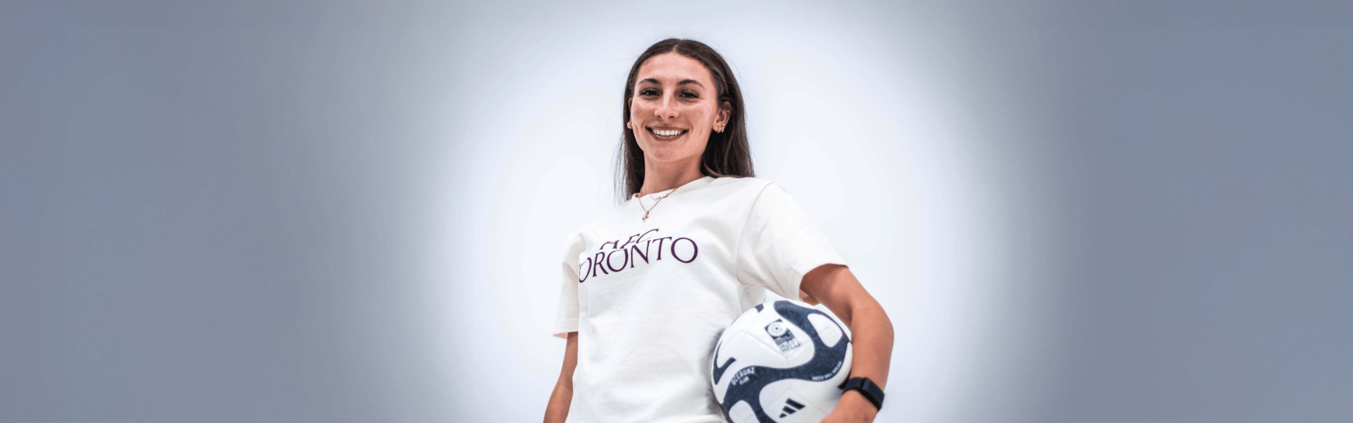 A woman with long brown hair stands smiling with a blue and white soccer ball under one arm while wearing a white t-shirt with a maroon AFC Toronto logo on the front.