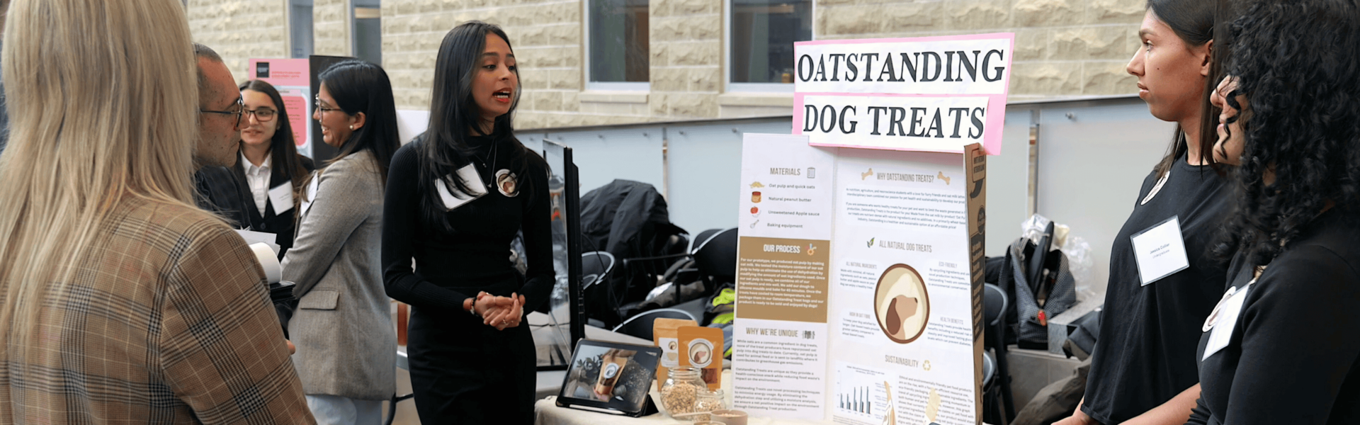 Student presenter stands in front of a display entiteld 'Oatstanding Dog Treats' as observers watch. Display contains food samples of their project