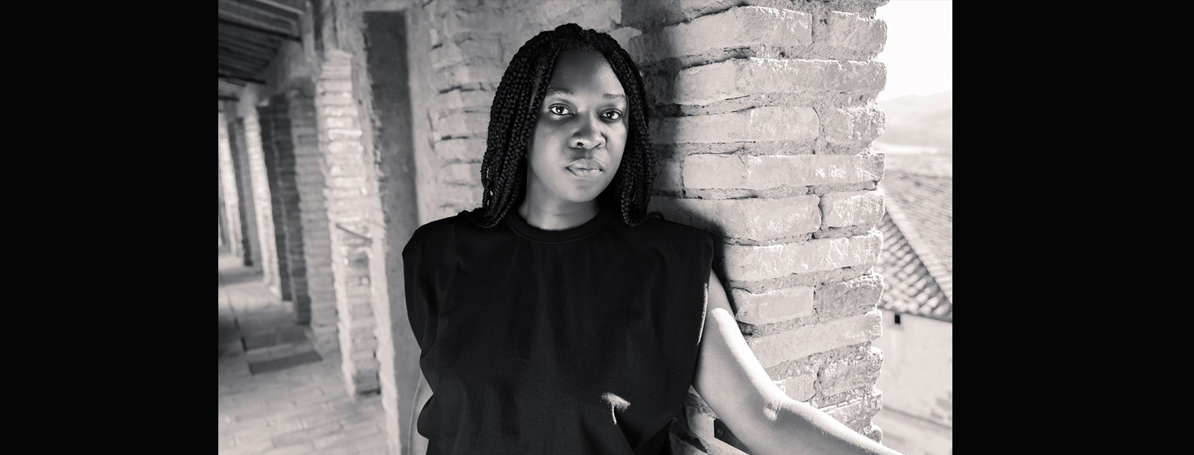 A person with long, black braids wearing a black sleeveless t-shirt stands against a brick wall with one arm extended on a ledge.