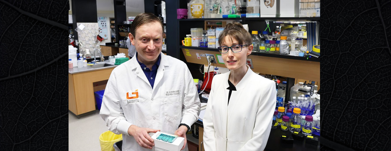 Two researchers in white lab coats face the camera smiling and one holds a blood analysis device.