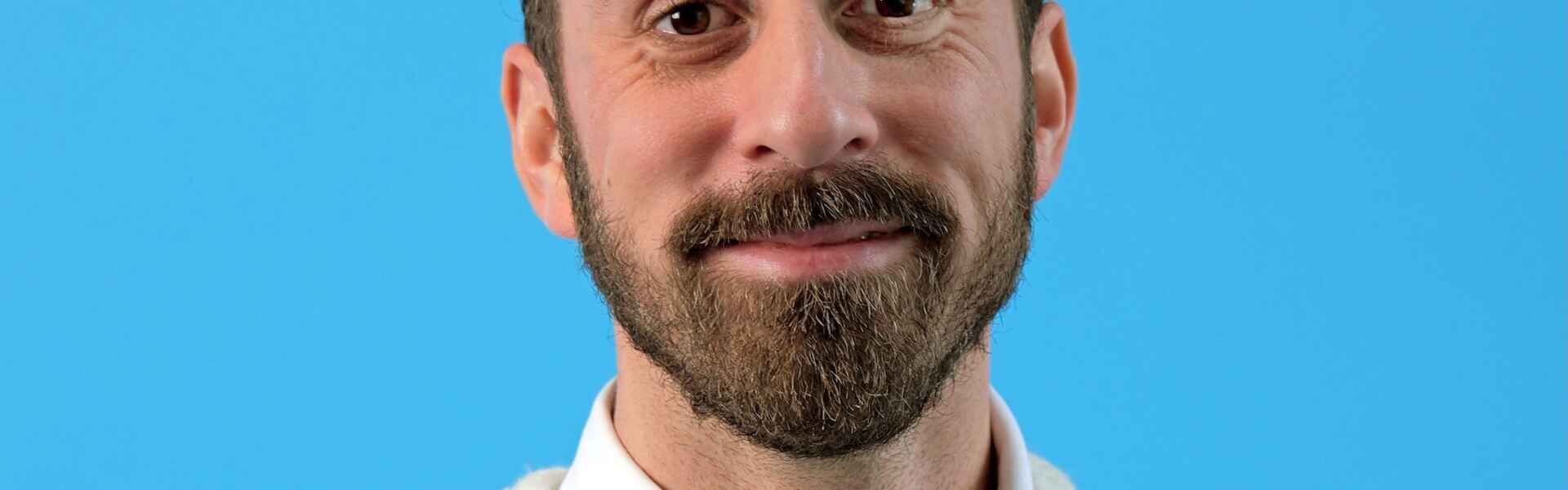 A person with short brown hair and a brown beard wearing a white collared shirt under a beige sweater smiles into the camera in front of a blue background