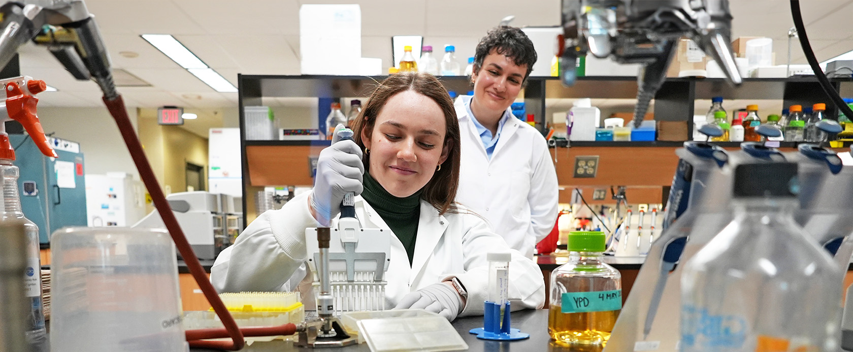 A person in a lab coat uses a multichannel pipette in a lab as another person watches and smiles