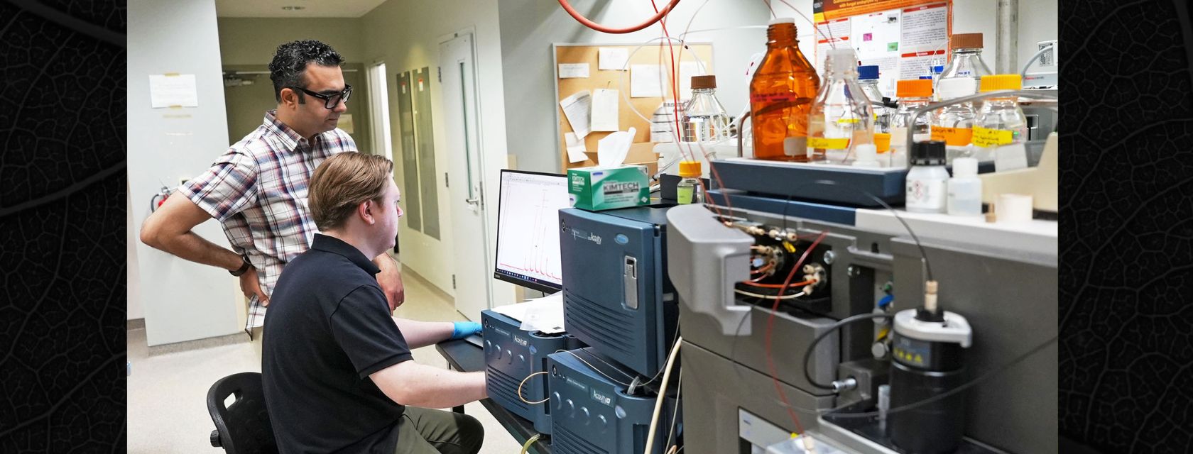 Two people with short brown hair in a lab, one seated at a computer and wearing plastic gloves while one looks on amidst various lab equipment.