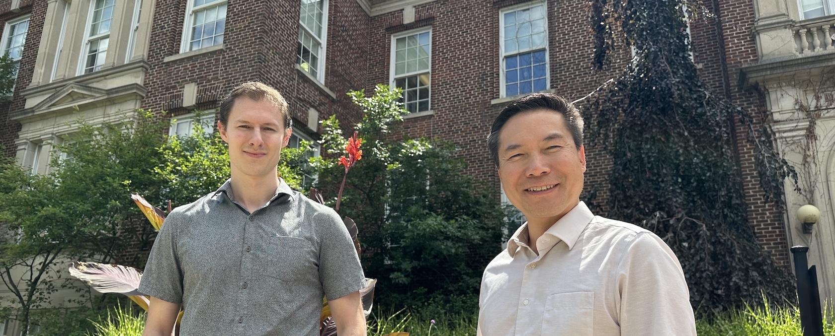 Two people, one in a blue shirt and the other in white stand smiling in front of a brown brick building with some greenery in the background