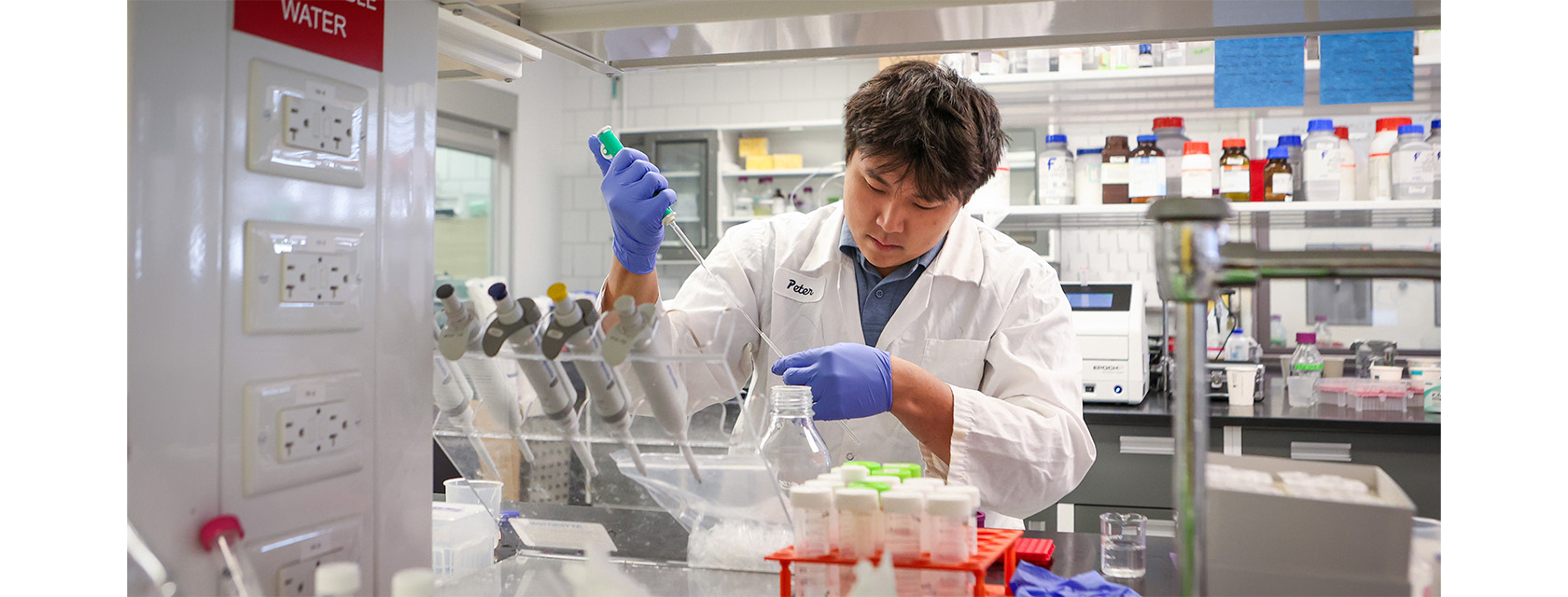 person in a lab coat uses a long pipette to add a clear liquid to a dish in a chemistry lab