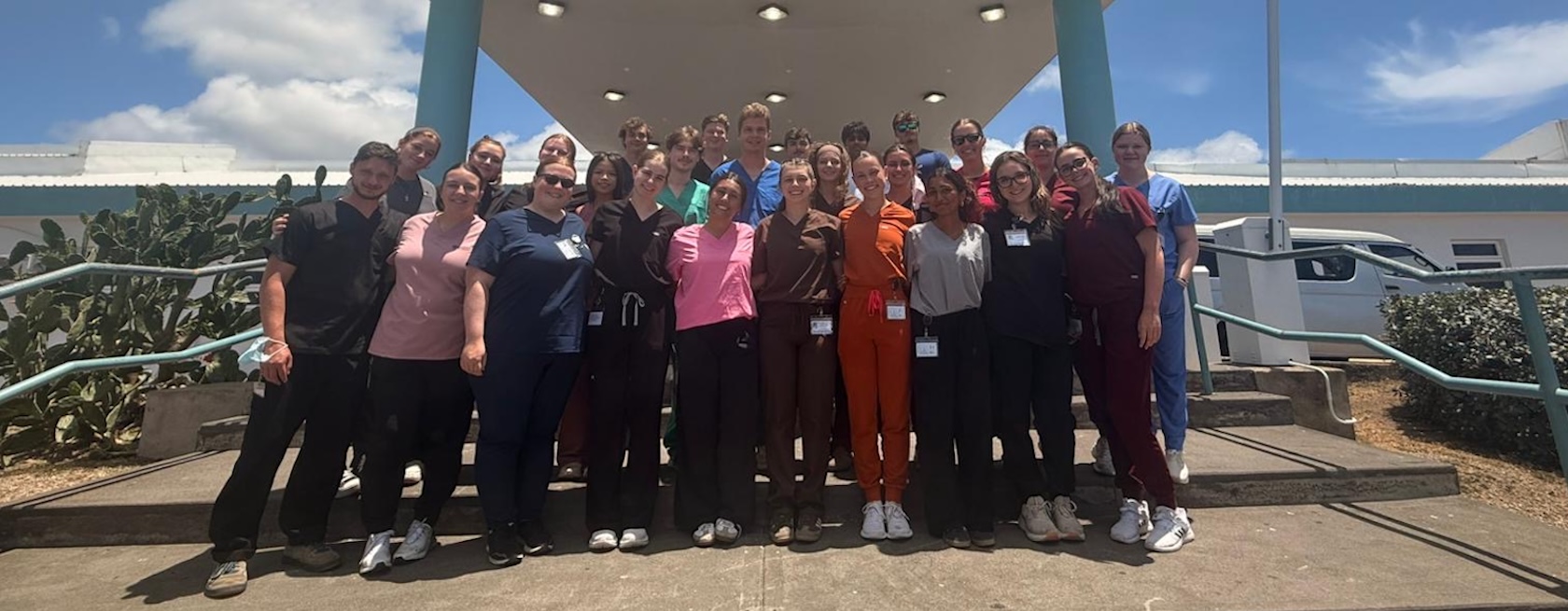 A group of students in medical scrubs of all colours stand together in a group shot outside a hospital in the Caribbean.